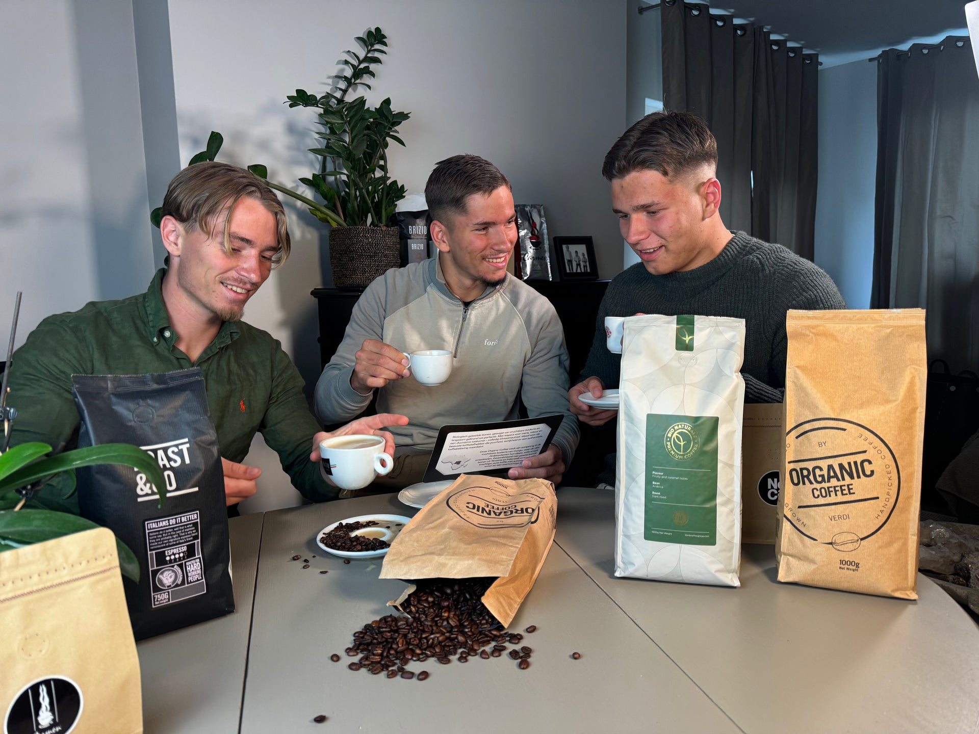 Drie jonge mannen drinken koffie en bespreken iets aan tafel, omringd door zakken specialty koffiebonen en gemorste koffiebonen op de tafel.
