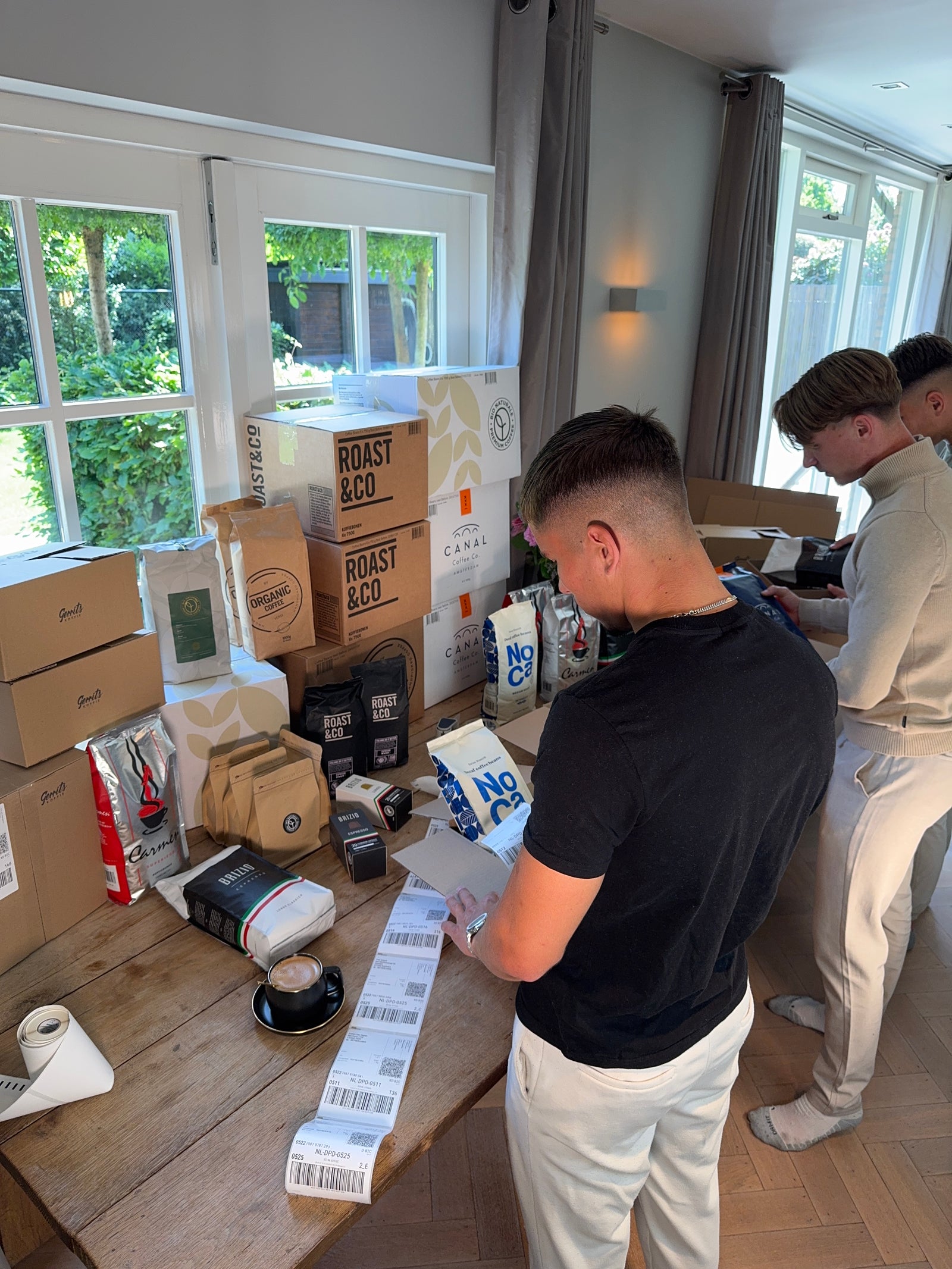 Twee jonge mannen verpakken diverse dozen en koffiezakken aan een houten tafel vol met Roast & Co, Canal Coffee en NoCa producten, met een raam en groene tuin op de achtergrond.
